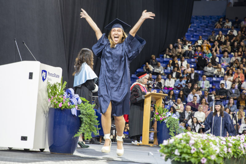 Student wearing cap and gown jumps for joy crossing the stage at commencement