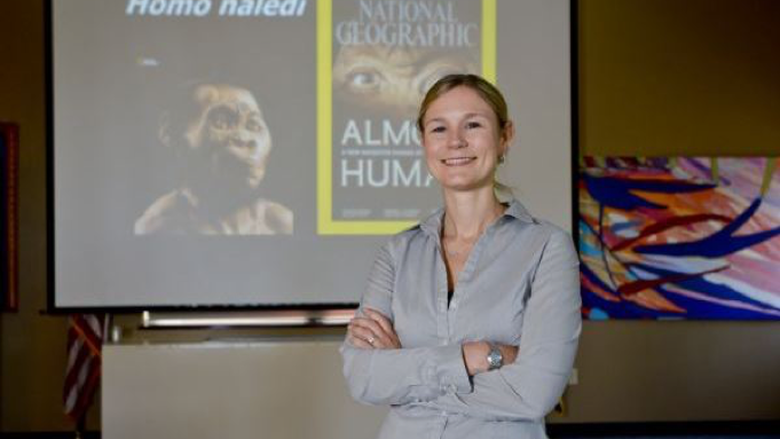 Dr. Heather Garvin poses in front of a screen, projected with imagine of Home naledi.