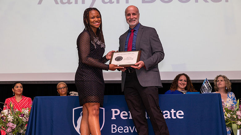 Student Aaliyah Booker stands with Dr. Chris Rizzo on a stage in front of a table with the Penn State Beaver logo as she accepts her award