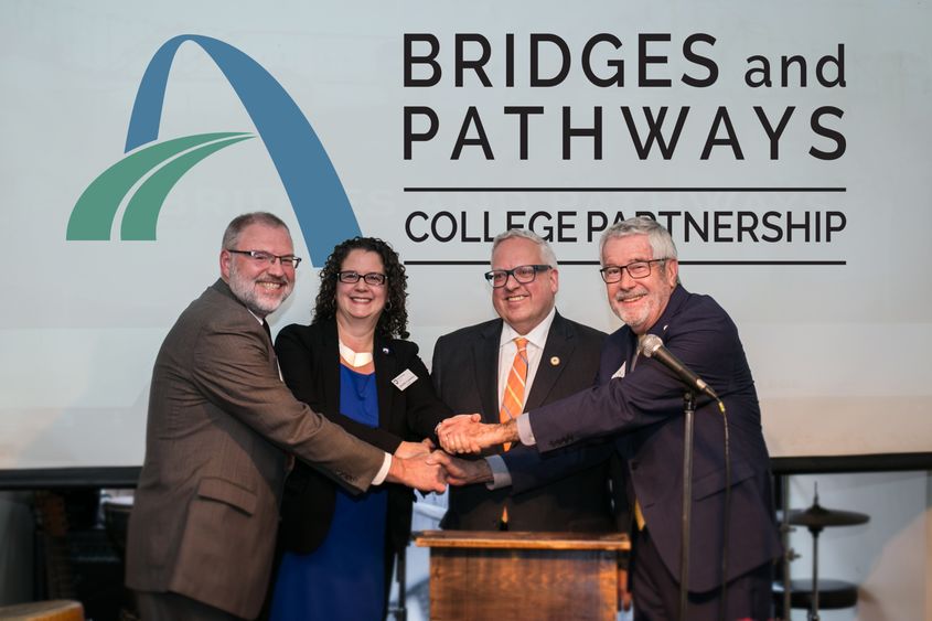 Four college presidents shake hands on stage.
