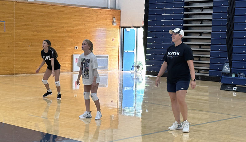 A female coach wearing navy blue and a ball cap stands to the right of the photo in a gymnasium as female volleyball players prepare to make a play to her left.
