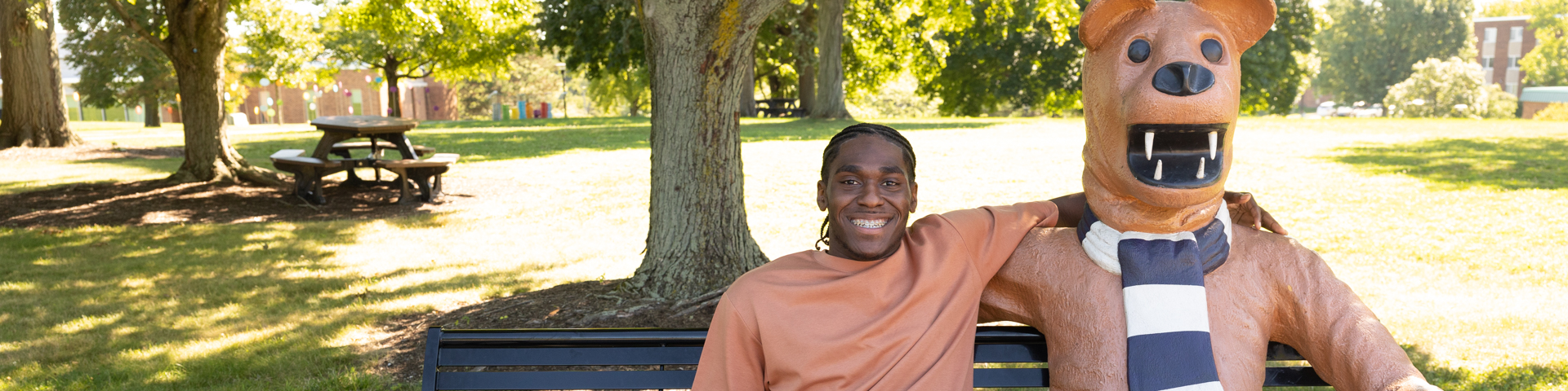 A male student sits on the Lion Bench next to the statue of the Nittany Lion mascot.