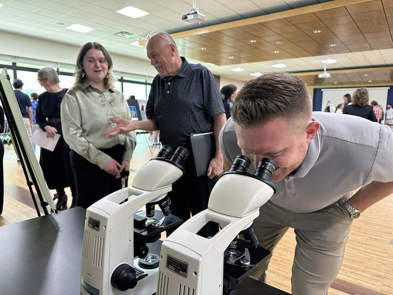 A man looks through a microscope while a student talks to another man