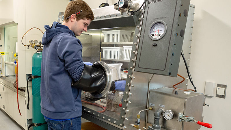 A student is working in front of a glass box. Rubber gloves, that are built into the box, extend up to his elbows as he reaches into the chamber.