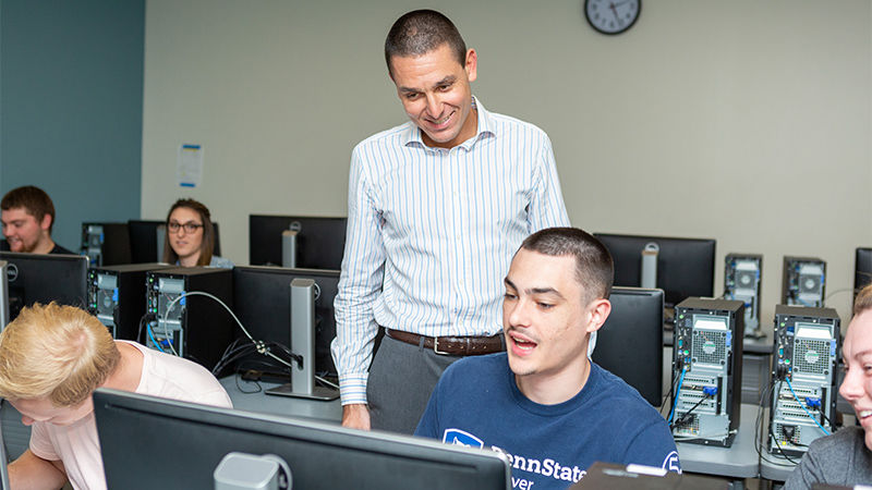 The professor, wearing a white dress shirt with blue vertical stripes, stands smiling behind a student seated at a computer.