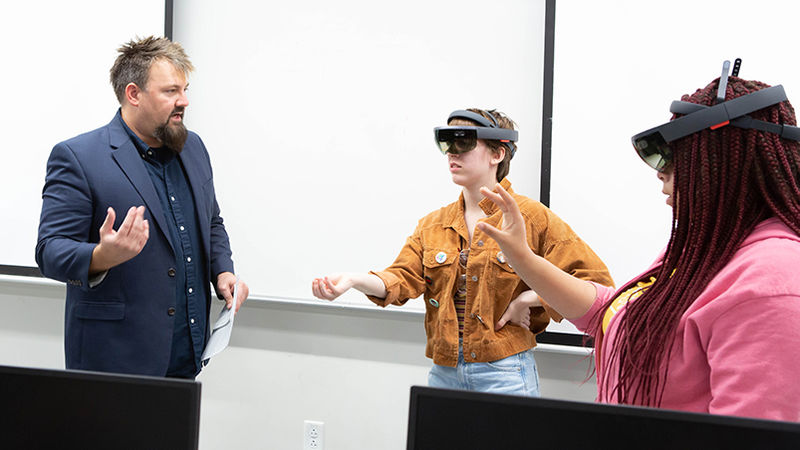 Kevin Bennett shows two female students how to use virtual reality headsets.