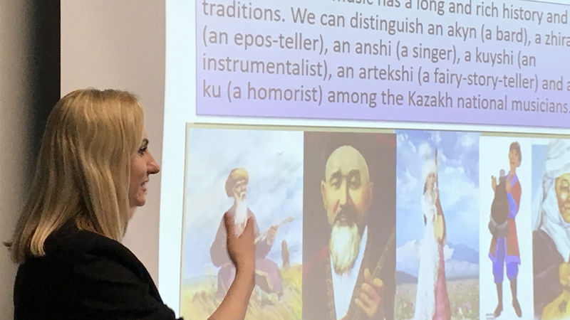 Professor from Kazakhstan talks to Penn State Beaver class A blonde woman wearing a dark jacket stands to the left of a screen displaying information about Kazakhstan.