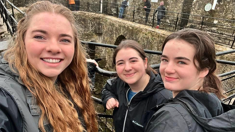 Three female students stand in the courtyard area of Blarney Castle and Gardens in Ireland. It looks to be cloudy and cool. Everyone is wearing coats.