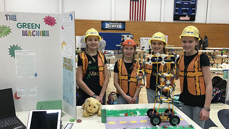 Four girls stand in front of a table wearing bright construction vests and yellow construction hats.