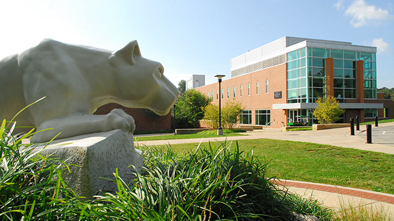 A Nittany Lion statue looms in the left foreground of the photo while the Ross Administration building can be seen in the background on a sunny day.