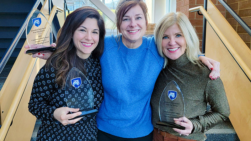 The three award winners stand in a stairwell holding glass awards.