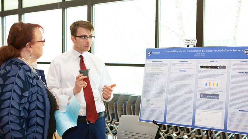 A male student wearing a white dress shirt and red tie explains his research to a professor in front of a poster board.