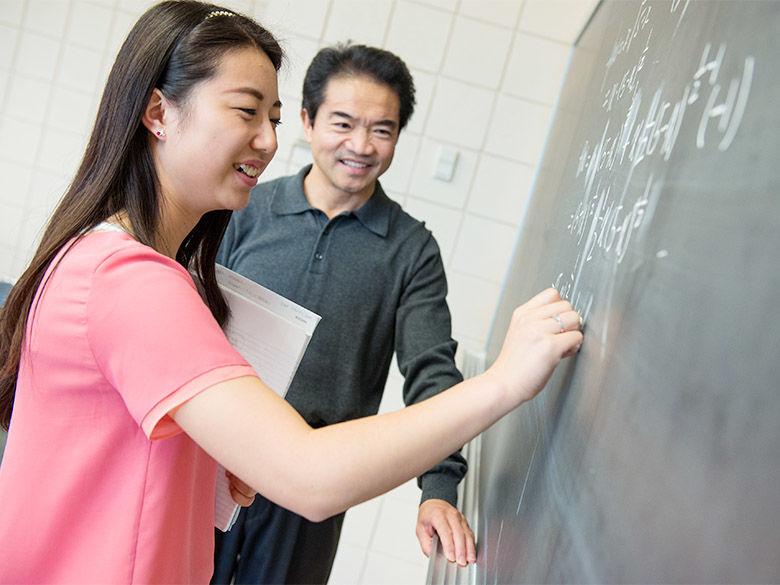 A professor helps a student do math problems at a chalkboard.
