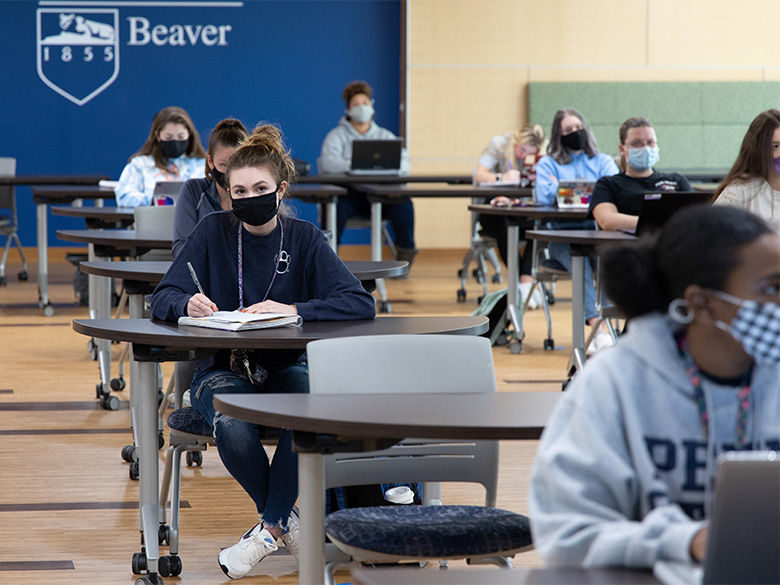 Students wearing masks take notes during class while sitting at widely spaced desks.