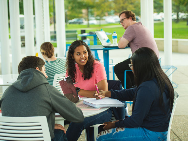 Several students sit at tables in an outside pavilion