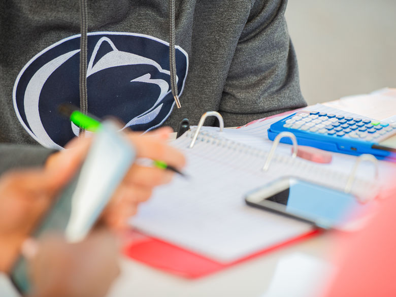 Closeup of notebooks, calculator, phones on a table. A student is wearing a shirt with the Penn State athletics logo.