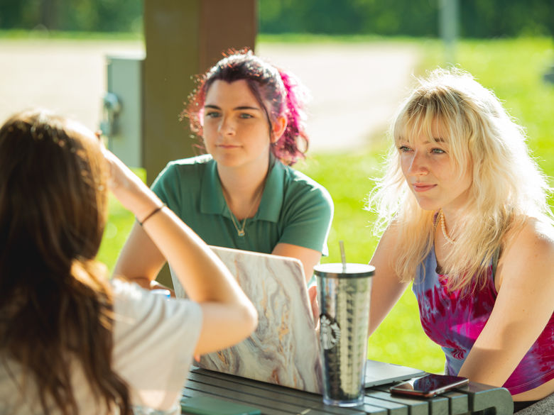 Three female students sit outside at a picnic table with their laptops open.