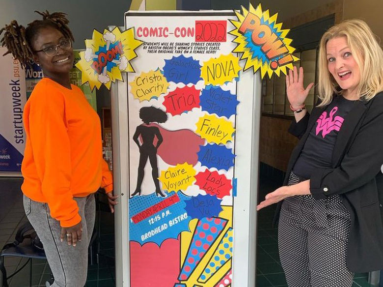 A student and a professor stand next to a poster advertising Comic-Con 2022.