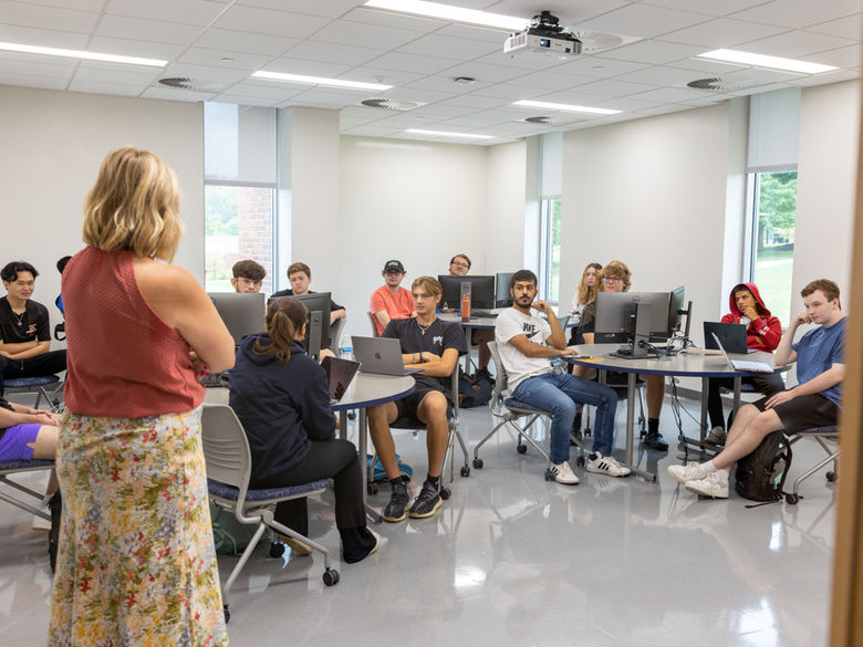 a female professor dressed in a skirt stands with her back to the camera while teaching a group of students sitting at round tables