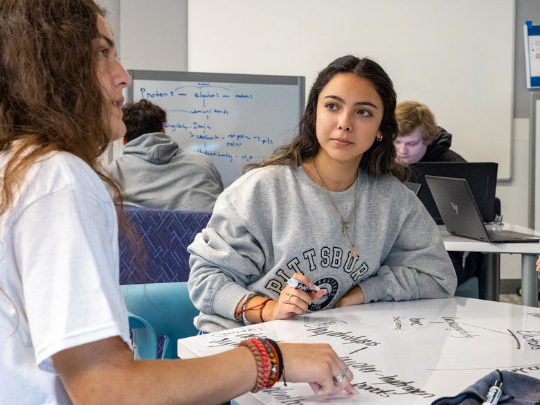 Two female students write on a whiteboard table while solving a problem.
