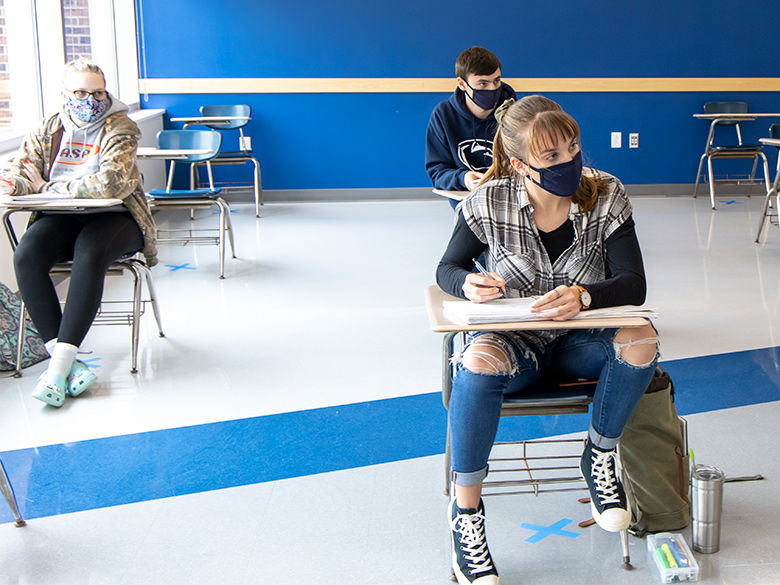 Three students wearing masks sit at desks in a classroom. All are paying attention to a lecture.