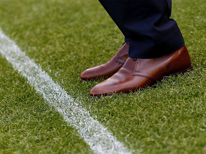 A close-up view of a person's legs and feet dressed in dark slacks and brown wing-tip shoes. The person is standing near a yard line on a football field.