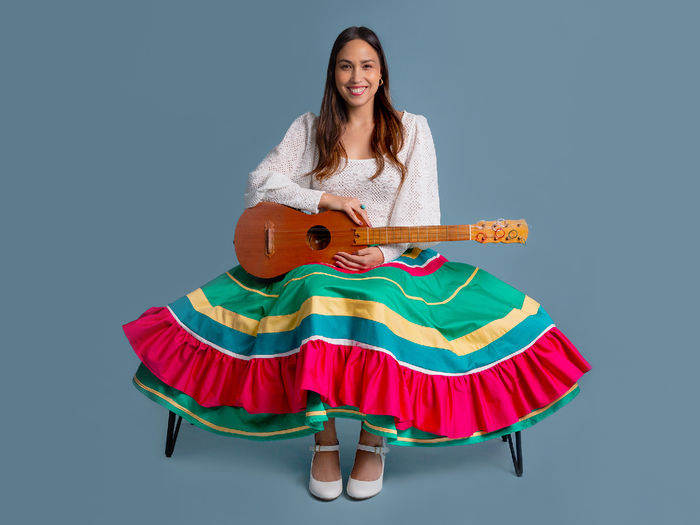 A smiling Sonia de los Santos wears a colorful green and red skirt and white shirt, and holds a guitar in her lap.