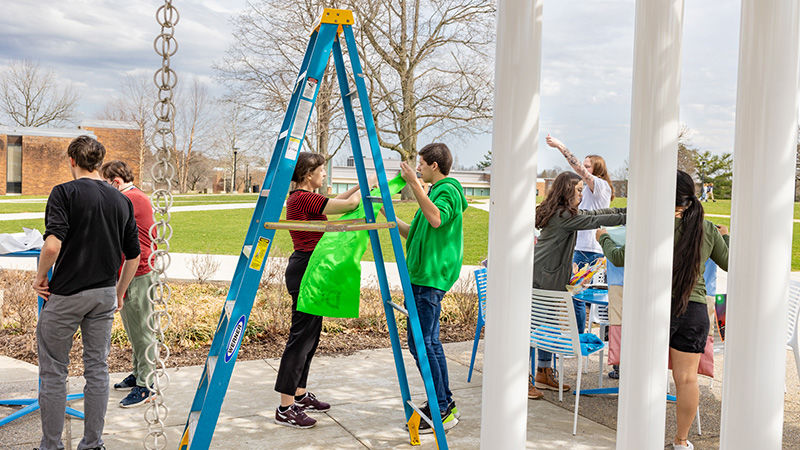 A female teacher, framed by a tall step ladder, helps a male student in a bright green sweatshirt, prepare to hang a flag.