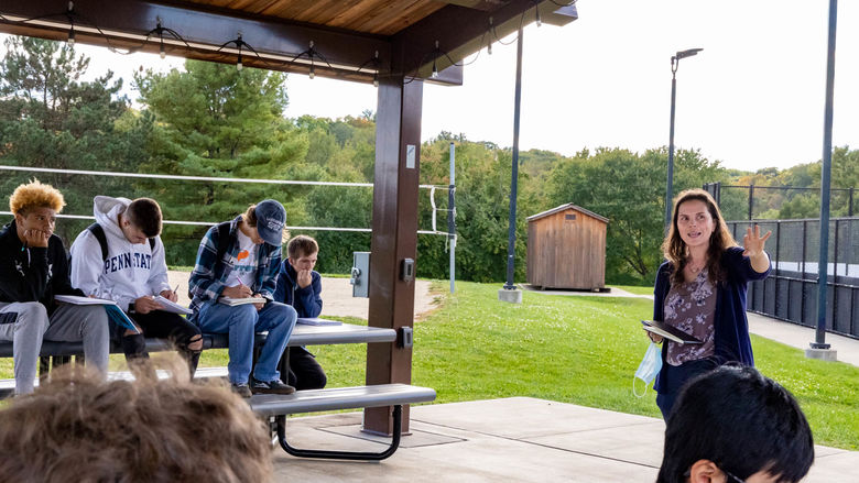A group of students sit under a pavilion with sketchbooks to the left of the photo while to the right a female professor gestures