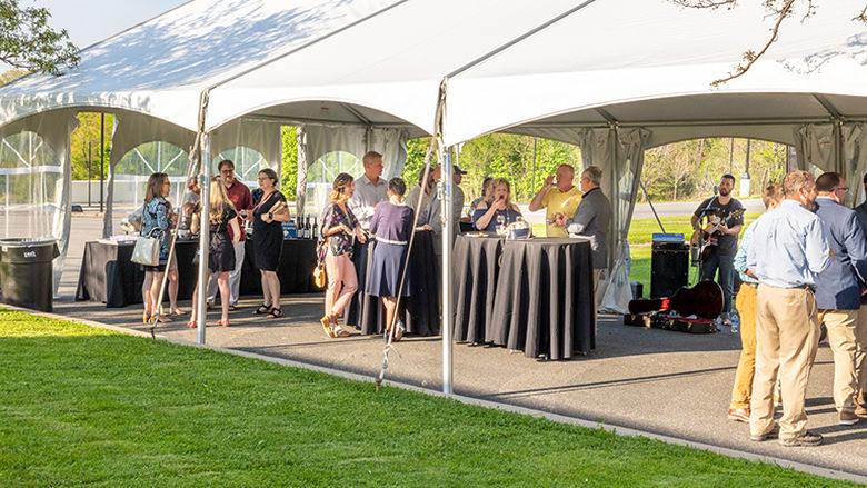 People gather under a white tent on a green lawn on a sunny evening. There are high-top tables under the tent.