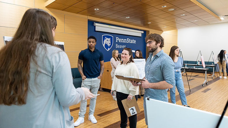 Students and library staff members discuss a student research project. The back wall is blue and displays the Penn State Beaver logo in white.