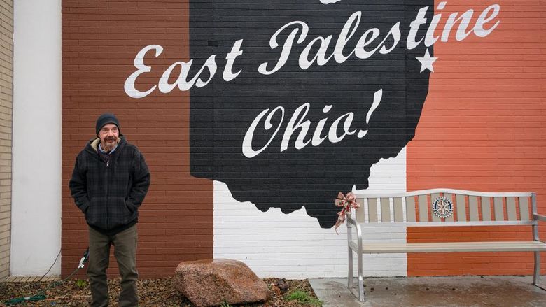 A man wearing a dark winter coat and a knit hat stands in front of a mural that says East Palestine, Ohio.