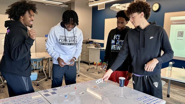 A group of four students stand around a whiteboard table with a custom-made board game on it