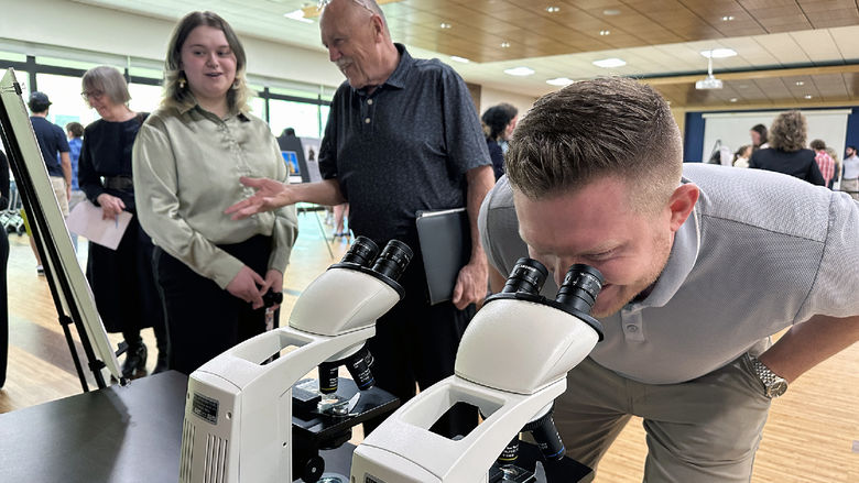 A man looks through a microscope while a student talks to another man