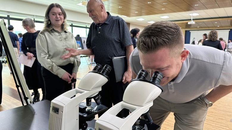 A man looks through a microscope while a student talks to another man