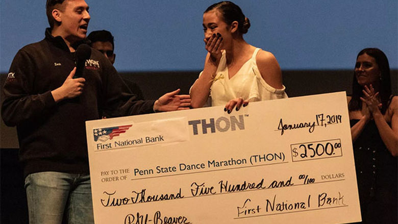 Dancer Hailey Burbage stands on stage, covers her mouth with her hand and reacts with emotion as she is presented a large check for winning the THON Showcase.