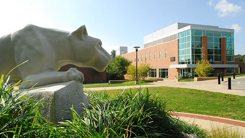 A statue of the Penn State Nittany Lion looms in the left foreground of the photo while the Ross Administration is seen in the background on a sunny day.