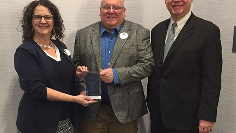 Scott Noxon wears a blue shirt and brown jacket as he accepts a glass award. Standing left is Chancellor Dr. Jenifer Cushman and to the right of the photo is Advisory Board President Scott Cunningham.