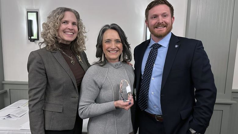 A photo of three people, and the woman in the center is holding a crystal award