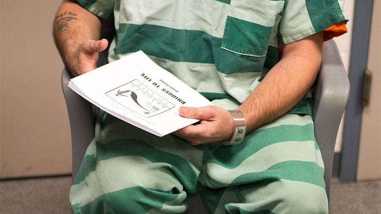 An inmate dressed in a prison jumpsuit holds a book that says Bridges to Life on the cover.
