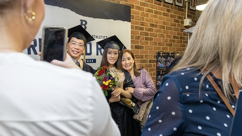 In the foreground someone takes a picture of Cindy in her cap and gown with her parents. Her father is also wearing a cap and gown, and her mother is in a mauve sweater