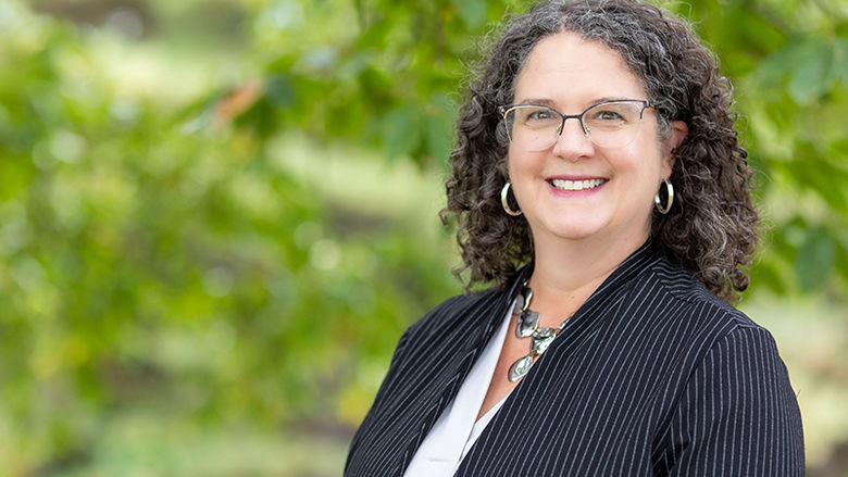 A headshot of Jenifer Cushman standing outdoor in front of a wooded area. She is wearing a dark blazer and white shirt. 