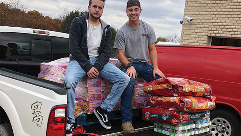 Two students sit on cases of pet food in a pickup truck.