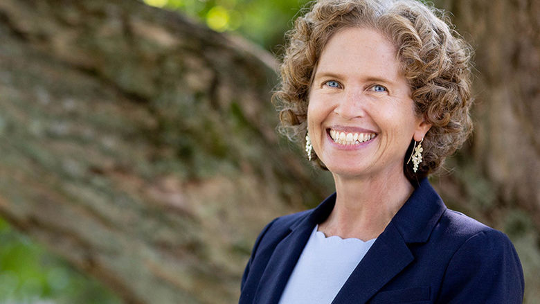 McDougall is pictured outdoors with trees in the background. She is smiling and wearing a light blue shirt and navy blue blazer.