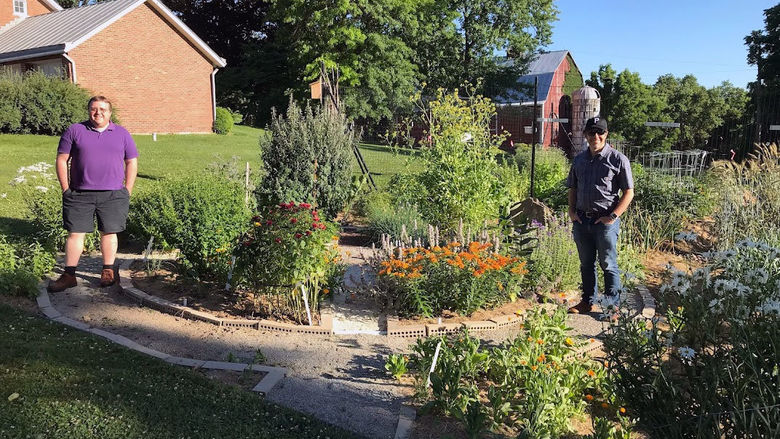 Two men stand beside a collection of plants in the sun