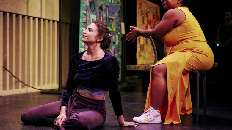 A white woman sits straight on a floor stage next to a Black woman sitting in a chair.