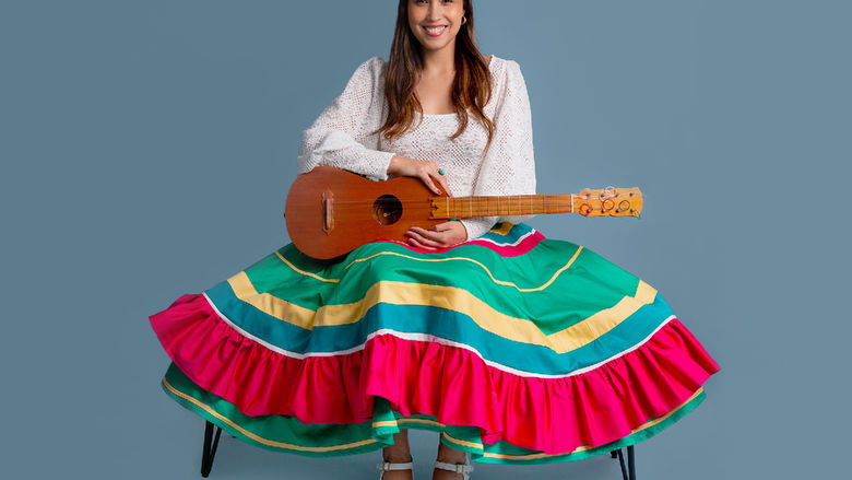 A smiling Sonia de los Santos wears a colorful green and red skirt and white shirt, and holds a guitar in her lap.