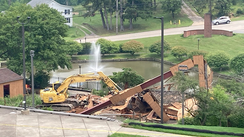 A piece of construction equipment sits in front of the remnants of a building. There is a pond with a fountain in the background.