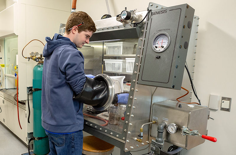 A student is working in front of a glass box. Rubber gloves, that are built into the box, extend up to his elbows as he reaches into the chamber.