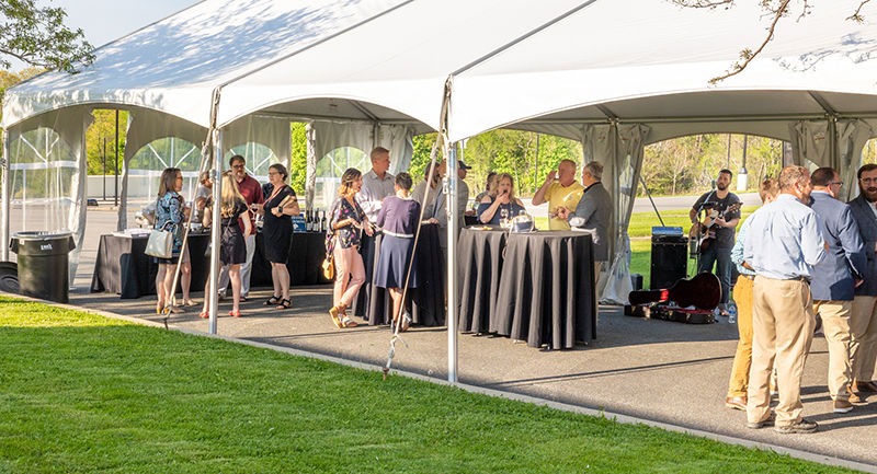 People gather under a white tent on a green lawn on a sunny evening. There are high-top tables under the tent.